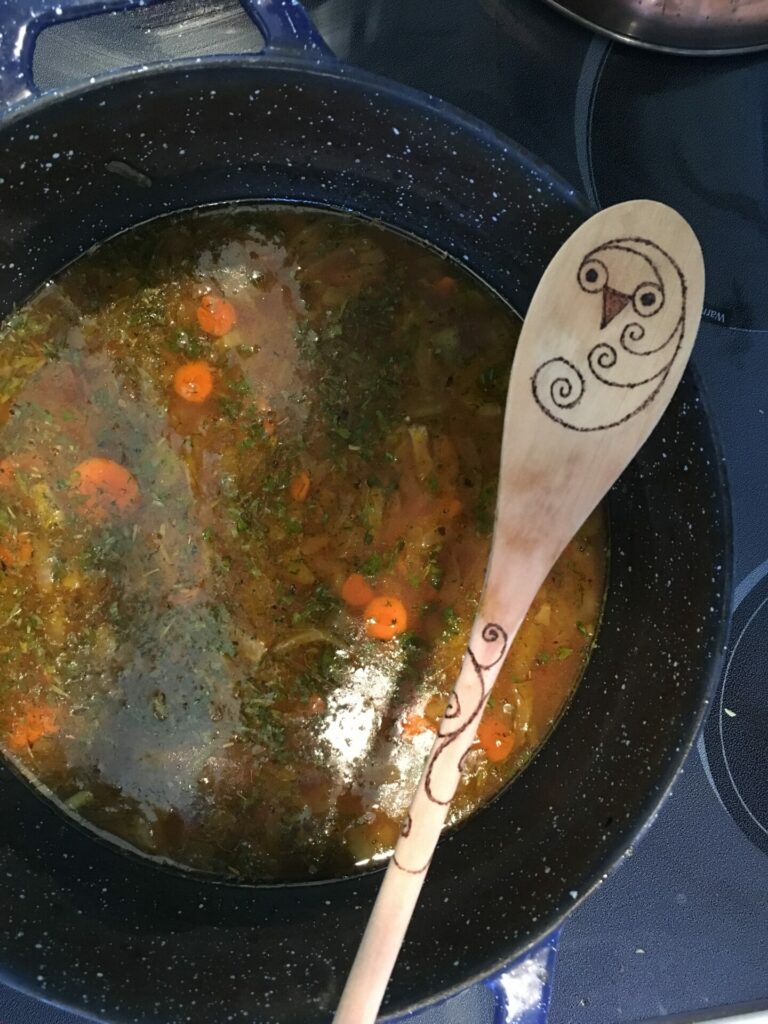 A wooden spoon with an owl design rests on the edge of a pot of soup. The spoon is part of a spiritual ritual.